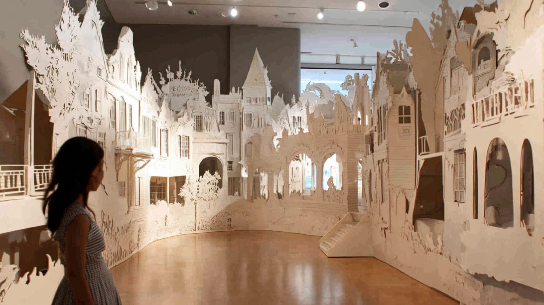 A woman observes a detailed cardboard cutout installation resembling an intricate town scene in a museum